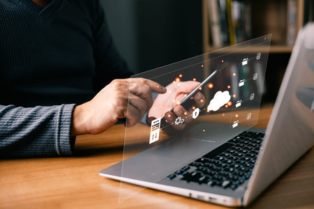 A person interacts with a smartphone beside a laptop using digital holographic icons representing...