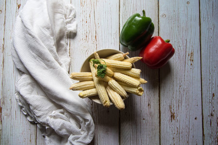 Top view of fresh baby corns in a bowl along with condiments on a background
