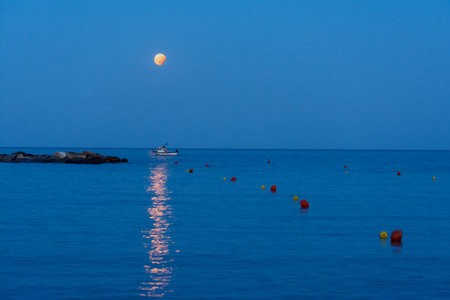 partial Moon eclipses due to the earth, while the moon rises on the sea during a summer evening, while a small fishing boat crosses, italyの写真素材