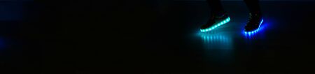Fashionable sneakers with neon LED lighting on the legs of a girl with blue and azure colors in the dark .Different colors of neon lights soles sneakers are present.Panorama view.の写真素材