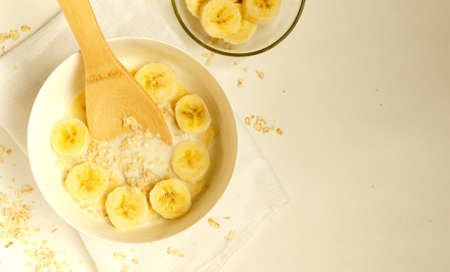 Oatmeal porridge with banana slices with a wooden spoon on white background, copyspace.Vegetarian healthy breakfast.の写真素材