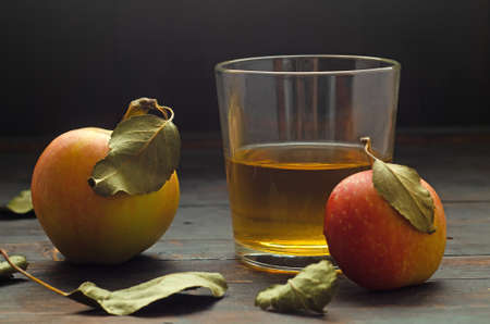 A glass with apple cider vinegar and two apples with leaves on wooden background.の写真素材