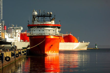 Industrial port scene with a red offshore supply vessel and an LNG tanker moored in the harbor....