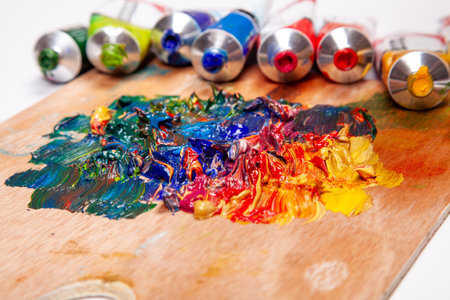 Vibrant oil paints mixed on a wooden artist's palette with tubes of color in the background. A...