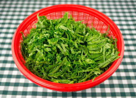 Freshly chopped raw mustard leaves in a red strainer bowl, against a green and white checkered tableclothの写真素材