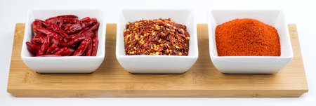 Chili peppers, flakes and cayenne powder in white bowls on a wooden tray against a white backgroundの写真素材