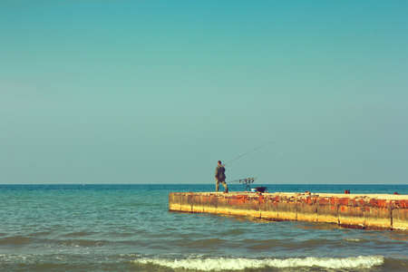 The man is a fisherman with a fishing rod on the breakwater, it is going to catch fish in the sea. Warm sunny summer day. Horizontal landscapeの写真素材