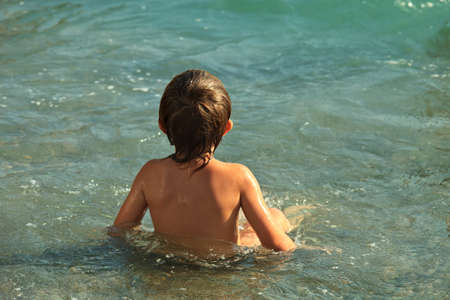 Boy about five years old to swim and sunbathe. Sitting in pure water. A child sits with his back to the camera. He looks into the distance. The sun is shining. Clear water glistens in the sun. Wet hair clung to his shouldersの写真素材