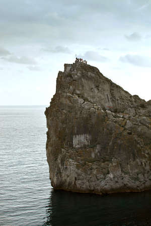 A rock in the sea. On top of a rock climbed a group of tourists people. People beat nature. Extreme trip. Travel in the mountains. Mountain stands in the water against a blue sky at sunset. The sea is calm, no waves. Seascape.の写真素材