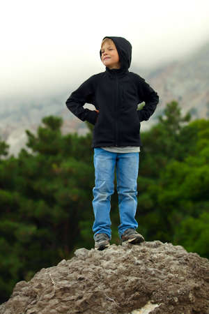 Boy teenager tourist standing on a rock and smiling. Man happy hike in the mountains. Against the background is seen the mountain and the forest, misty landscape and overcast sky. Coniferous forest, green trees. Boy is putting his hands on his hips. Teenaの写真素材