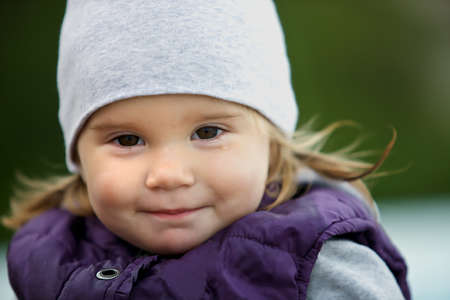 Cute sad little girl smiling. Baby wearing a warm hat and jacket. From under the cap can be seen long hair. The little girl smiles and grimaces. It's cold and cloudy. Photo close-up shot. Portrait.の写真素材