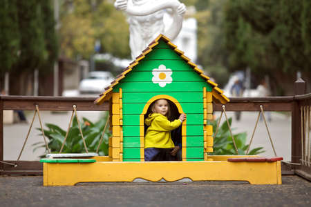 In Park is a small colorful games. House made of wood, brightly painted. In the house painted flower. From the door the house looks and smiling little girl in a warm fleece suit. The little girl looks at the camera and plays the game. Outdoor warm weatherの写真素材