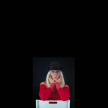 Minimalist Portrait with coys pace. of a mature woman with long white hair dressed in red sweater wearing a gray and black hat sitting on a backward white chair against a black background.の写真素材