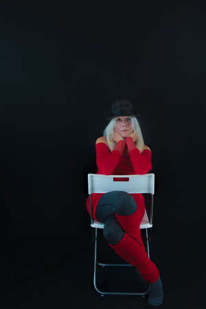 Copyspace available on black backdrop in portrait studio shot of a mature woman dressed in red sweater and leg warmers, gray leggins and socks and gray and black hat, and long white hair.  Hands supporting chin with elbows resting on a white chair back, legs crossed through chair back openingの写真素材