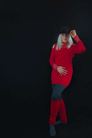 Portrait of a mature wearing gray and black hat on top of white hair, red sweater and leg warmers and gray leggings.  Posed in a minimalist style against black backgroundの写真素材
