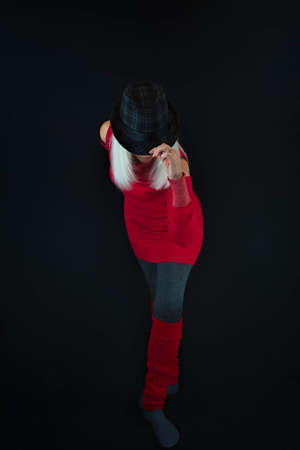 A mature woman wearing gray leggings and socks, red sweater and leg warmers and a black and gray hat on top of a head of white hair, taking a bow in this studio shot self portrait.の写真素材