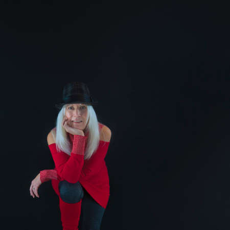 Portrait of mature woman with long white hair dressed in gray leggingsand socks, red leg warmers and sweater, wearing a gray and black hat against a black background with copyspace.の写真素材