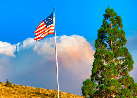 A controlled fire sends up a smoke cloud behind a hill in Lakeview, Oregon where the United States Flag flies from it's poleの写真素材