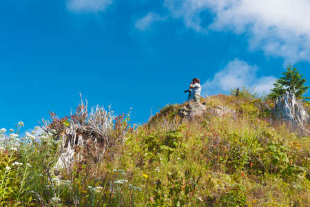 Photographer high on a ridge looks through binoculars, his camers mounted to a tripod stand next to him.の写真素材