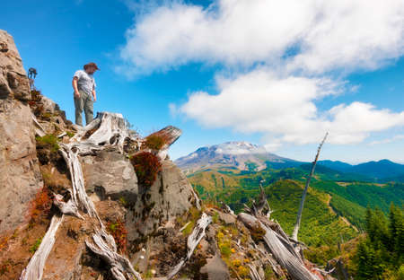 A hiker/photographer looks down from on top of Castle Peak, his tripod and camera close by with Mt. St. Helens in the background in Gifford Pinchot National Forest.の写真素材