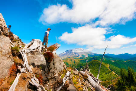 A hiker/photographer looks down from on top of Castle Peak, his tripod and camera close by with Mt. St. Helens in the background in Gifford Pinchot National Forest.の写真素材