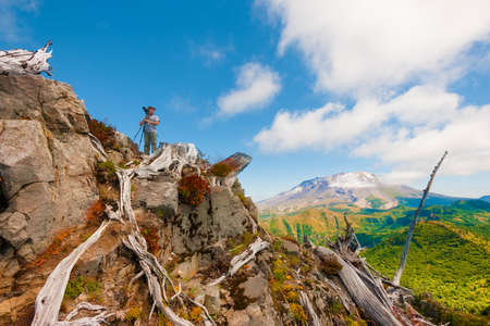 A hiker/photographer looks away from his tripod and camera, on top of Castle Peak with with Mt. St. Helens in the background in Gifford Pinchot National Forest.の写真素材