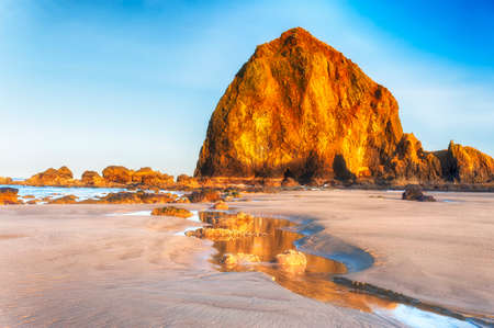 The rising sun cast golden rays lighting a watery path that leads to Haystack Rock.  accented by wispy clouds and blue skies. during low tide on Cannon Beach, Oregon.の写真素材