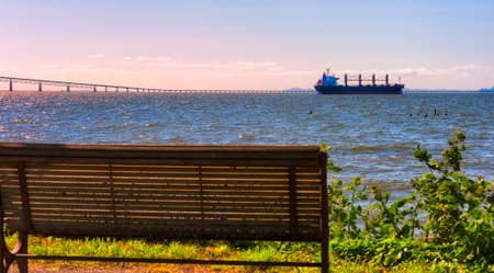 A bench awaits for someone to sit and enjoy the ever changing mural along Astoria's Waterfront.  A freighter heads into the near setting sun, to pass under the four mile long Megler Bridge towards the Pacific Ocean.の写真素材