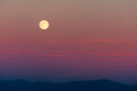 Harvest moon above the Cascade Mountain range.  The setting sun adds pastel colors to the sky while whispy clouds drift by.の写真素材