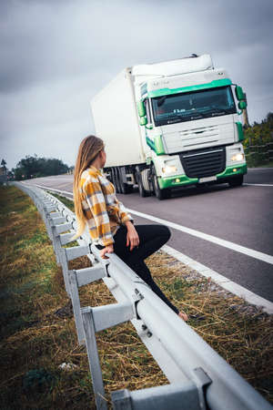 A girl sits by the road and waits for her transport. A truck is coming to meet the girl.