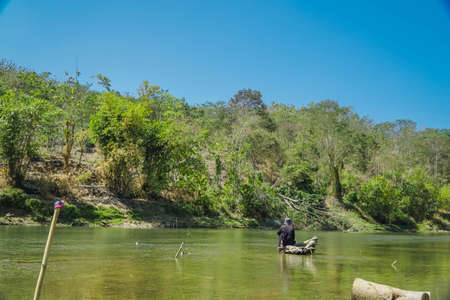 Old man is fishing in the middle of the river while sitting on a tree trunk