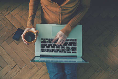 Laptop and coffee cup in girls hands sitting on a wooden floor
