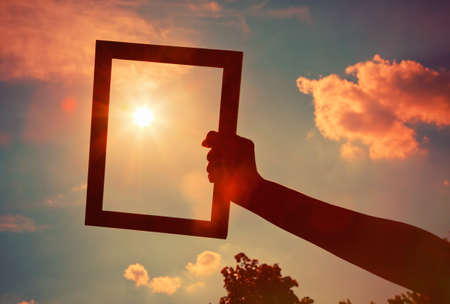 Hand holding a wooden frame on sunrise sky background. Care, safety, memory or painting concept.