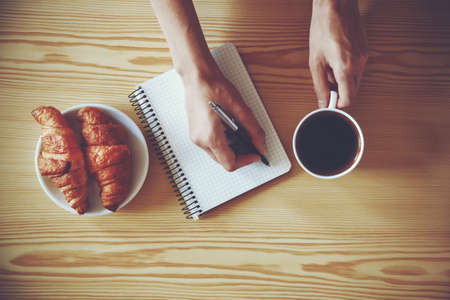 Hands with pen writing on notebook with morning coffee and croissant. View from aboveの写真素材