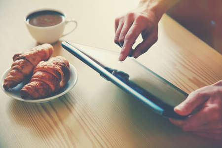 Hands holding digital tablet pc with morning coffee and croissant.