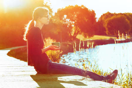 Smiling girl with cup of tea or coffee enjoying on summer shore of riverの写真素材
