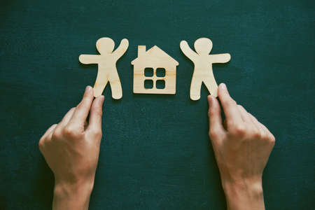 Hands holding little wooden men and house on blackboard background. Symbol of construction, sweet home conceptの写真素材
