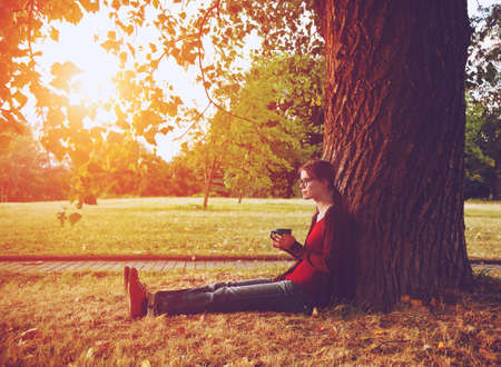 Smiling girl with cup of tea or coffee enjoying near park tree in morning sunlightの写真素材