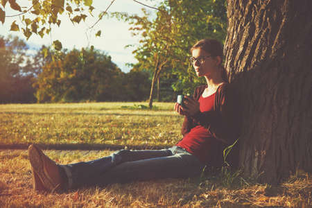 Smiling girl with cup of tea or coffee enjoying near park tree in morning sunlightの写真素材