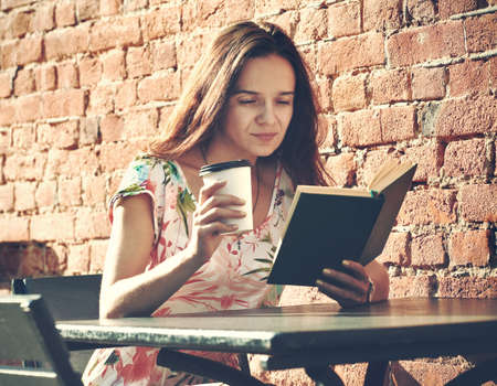 smiling girl sitting at summer terrace in street with morning coffee and reading book