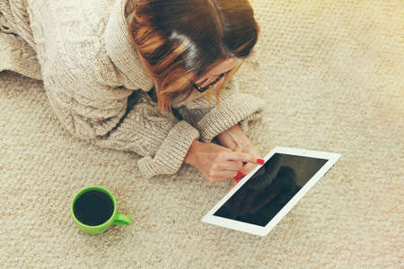 Woman lying on floor with digital tablet touching with finger and cup of morning coffee. View from aboveの写真素材