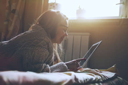 woman lying in bed with digital tablet touching with finger in morning sunlightの写真素材