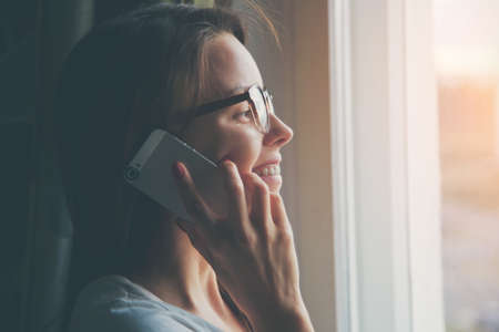 pretty woman talking on phone near window at homeの写真素材