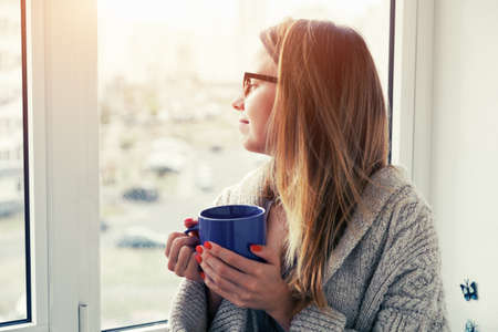 cheerful girl drinking coffee or tea in morning sunlight near windowの写真素材