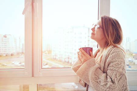 cheerful girl drinking coffee or tea in morning sunlight near windowの写真素材