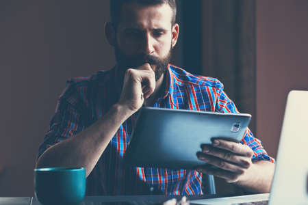 concentrated bearded man sitting with digital tablet and cup of coffeeの写真素材