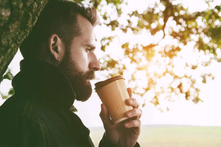Bearded man with paper cup of morning coffee walking in park