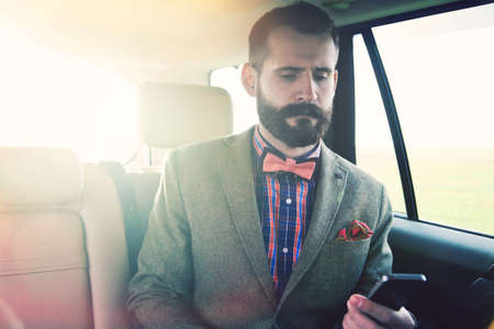 handsome bearded businessman sitting on back seat of car and touching phone