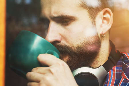 bearded man with headphones sitting with cup of morning coffee or tea near windowの写真素材