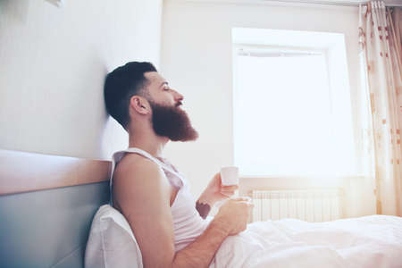 Bearded man in bed drinking morning espresso coffee in sunrise lightの写真素材